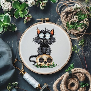 May include: A cross-stitch embroidery hoop featuring a black cat with orange ears and large eyes, perched atop a skull. The design is set against a white background, framed by a wooden hoop. The theme is Halloween.