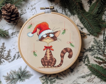 May include: Cross-stitch embroidery of a brown cat wearing a Santa hat and a red bow tie. The cat is in a wooden embroidery hoop, with hanging mistletoe accents. The background includes pine needles, pine cones, and a vintage paper.