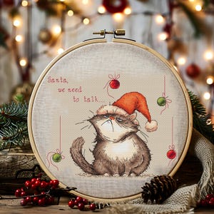 May include: A cross-stitch embroidery hoop featuring a grumpy cat wearing a Santa hat. The text reads "Santa, we need to talk..." with red and green ornaments. The cat is brown and white, with a stern expression. The hoop is set against a festive background.