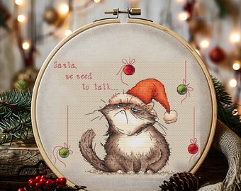 May include: A cross-stitch embroidery hoop featuring a grumpy cat wearing a Santa hat. The text reads "Santa, we need to talk..." with red and green ornaments. The cat is brown and white, with a stern expression. The hoop is set against a festive background.