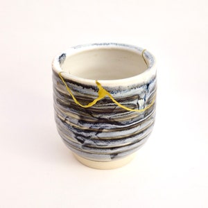 May include: A small, white ceramic cup with a blue and black glaze and gold accents. The cup has a slightly rounded shape and a wide base.