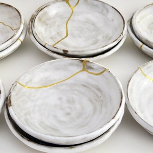 May include: Set of six white ceramic bowls with gold crackle glaze. The bowls are stacked in pairs and have a rustic, handmade look.
