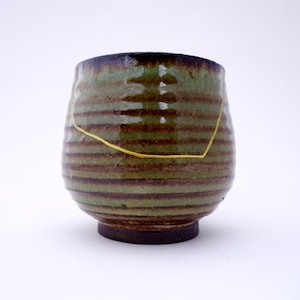 May include: A green and brown ceramic cup with a gold line across the middle. The cup has a textured, striped design.