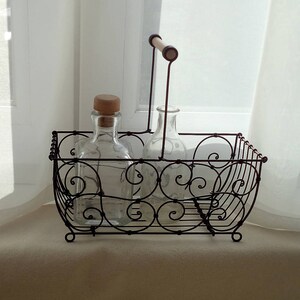 Könnte beinhalten: Ein Drahtkorb mit einem Holzgriff hält zwei klare Glasflaschen mit Korkverschlüssen. Der Korb ist mit einem geschwungenen Muster verziert.