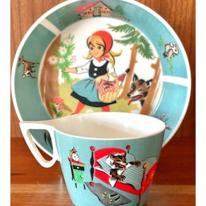May include: A vintage children's dish set, including a plate and cup, with a Little Red Riding Hood theme. The plate has a teal border and a central illustration of the character. The cup has a matching design with the wolf in bed. The set is on a wooden surface.