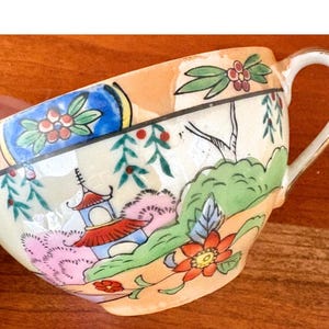 May include: A vintage teacup with a handle, featuring a colorful landscape design with a pagoda, flowers, and foliage. The cup has a cream-colored interior and a peach-colored exterior.