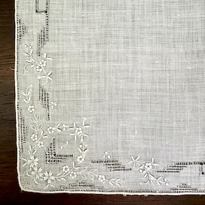 May include: A white linen handkerchief featuring intricate white floral embroidery and geometric cutwork patterns along the edges. The fabric has a visible woven texture, and the delicate needlework creates a decorative border design.