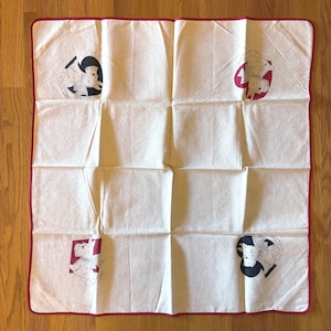 May include: A square off-white cotton cloth with a thin red border. The cloth features four printed illustrations of a terrier dog wearing a checkered bow, each placed within a different colored shape: a black spade, a red heart, a red square, and a black heart.