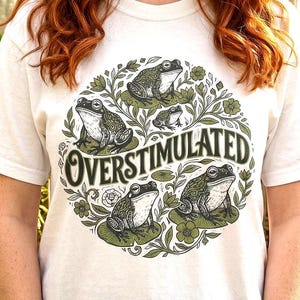 May include: White t-shirt featuring a circular design with several green frogs on lily pads, surrounded by green foliage. The text "OVERSTIMULATED" is in a stylized font. The shirt is made of a soft, light-colored material.