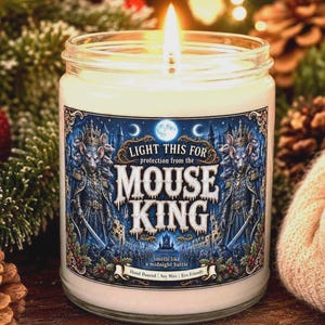 May include: A clear glass jar candle with a lit wick and white wax. The label features a blue illustration of a mouse king in armor with the text: LIGHT THIS FOR protection from the MOUSE KING, smells like a midnight battle, Hand Poured | Soy Wax | Eco Friendly.