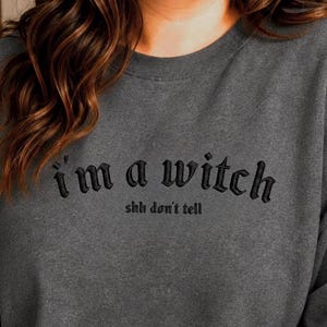 May include: A dark heather gray crewneck sweatshirt featuring black embroidered text on the chest that reads "I'm a witch" in a stylized font, with the smaller text "shh don't tell" positioned directly underneath in a simple sans-serif font.