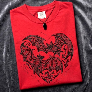 May include: A red t-shirt featuring a black heart-shaped bat design. The shirt has a crew neck and short sleeves. A black pendant necklace with a coffin-shaped charm rests on the shirt. The tag reads "Comfort Colors".
