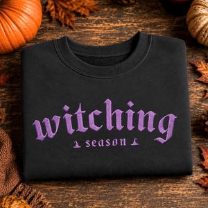 May include: A black crewneck sweatshirt featuring the word witching embroidered in purple thread in a gothic font, with the word season underneath in a smaller font, flanked by two small purple embroidered witch hat icons.