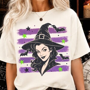 May include: An off-white t-shirt featuring a graphic print of a person wearing a black witch hat. The design includes horizontal purple brush strokes, black bats, and bright green stars surrounding the central figure with dark hair.