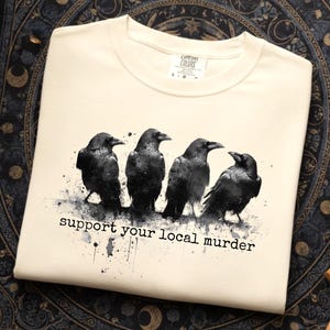 May include: Cream-colored t-shirt featuring a graphic of four black crows with a watercolor effect. The text "support your local murder" is printed below the birds. The shirt is folded and displayed on a patterned surface.
