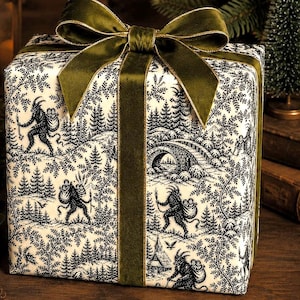 Pode incluir: Uma caixa de presente quadrada embrulhada em papel branco com uma estampa preta estilo toile, apresentando figuras com chifres em uma paisagem florestal. A caixa é amarrada com uma fita de veludo verde-oliva escuro formando um laço.