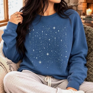 May include: A blue long-sleeve sweatshirt featuring a white graphic print of various sized stars and sparkling celestial points scattered across the front. The garment has a crew neckline and ribbed cuffs, providing a simple and comfortable fit.