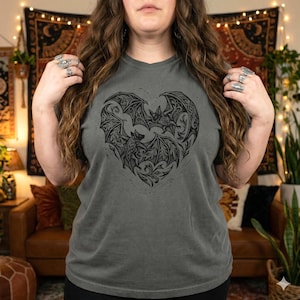 May include: Gray t-shirt featuring a black heart-shaped design with two bats. The bats have detailed wing patterns and are surrounded by floral elements. The shirt has a v-neck and a relaxed fit.