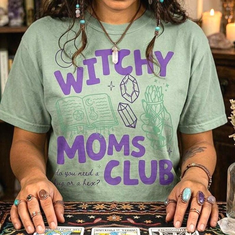 Op de afbeelding: Saliegroen t-shirt met de tekst "WITCHY MOMS CLUB" in paars. Het ontwerp bevat illustraties van een boek, kristallen en kruiden. De tekst "do you need a hug or a hex?" staat er ook op.