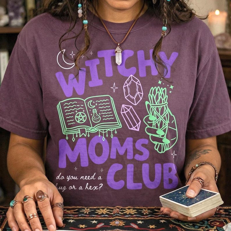 Op de afbeelding: Een paars t-shirt met de tekst "Witchy Moms Club" in paars en groen. Het shirt heeft illustraties van een halve maan, kristallen, een open boek en een bundel kruiden. De tekst "do you need a hug or a hex?" staat er ook op.