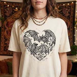 May include: Cream-colored t-shirt featuring a black heart-shaped design with two bats and ornate details. The bats are depicted with their wings spread, and the design has a woodcut style. The shirt has short sleeves and a crew neck.