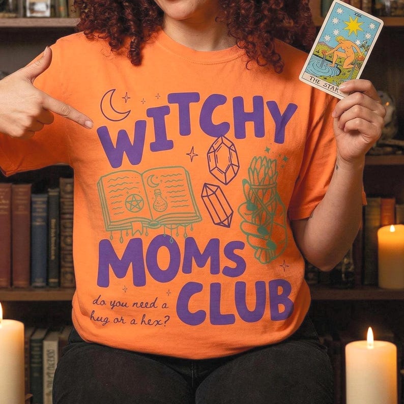 Op de afbeelding: Oranje t-shirt met de tekst "WITCHY MOMS CLUB" in paars, met illustraties van een boek, kristallen en kruiden. Het shirt bevat ook de zin "do you need a hug or a hex?" Een tarotkaart wordt in de hand gehouden.