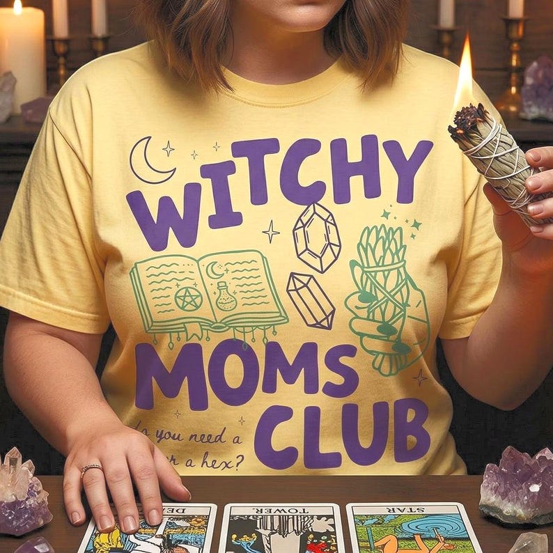 Op de afbeelding: Geel t-shirt met de tekst "WITCHY MOMS CLUB" in paars, met illustraties van kristallen, een boek en kruiden. De persoon draagt het t-shirt en houdt een brandend saliebundel vast.