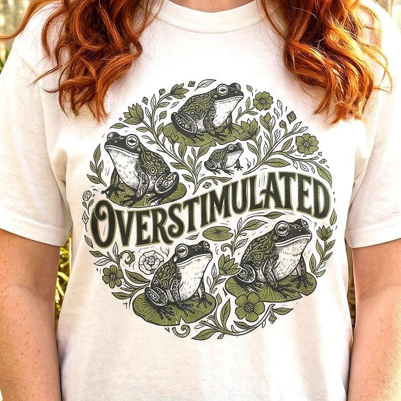 May include: White t-shirt featuring a circular design with several green frogs on lily pads, surrounded by green foliage. The text "OVERSTIMULATED" is in a stylized font. The shirt is made of a soft, light-colored material.
