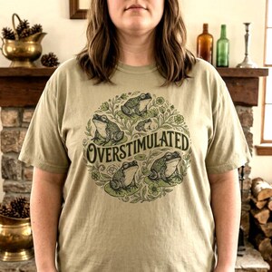 May include: A light olive green t-shirt featuring a circular design with several frogs and the word "OVERSTIMULATED" in a stylized font. The frogs are depicted in a cartoon style, surrounded by leaves and flowers.