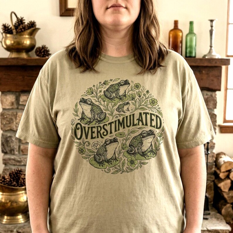 May include: A light olive green t-shirt featuring a circular design with several frogs and the word "OVERSTIMULATED" in a stylized font. The frogs are depicted in a cartoon style, surrounded by leaves and flowers.