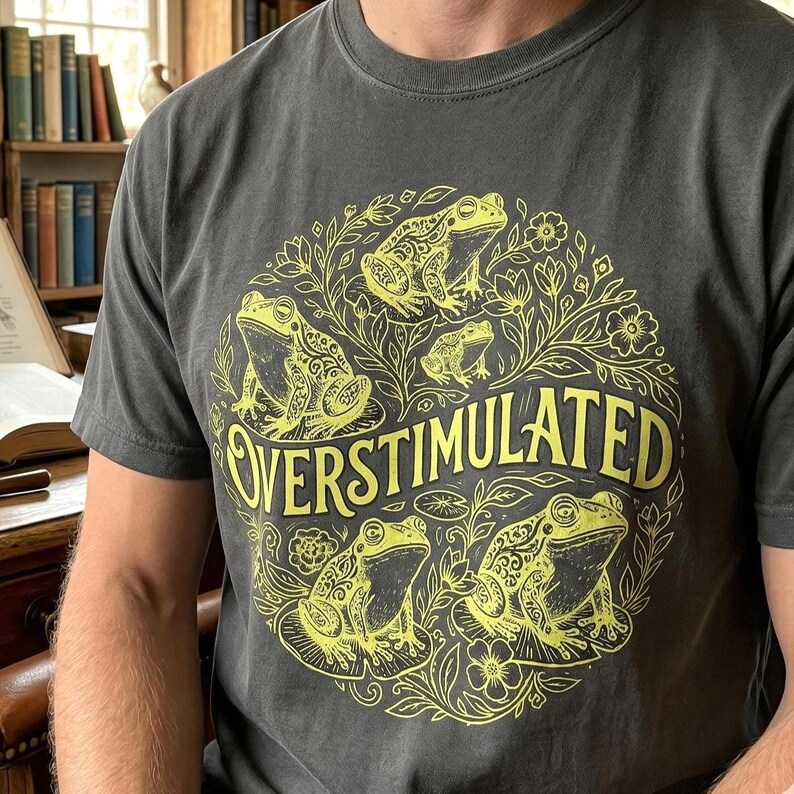 May include: Dark gray t-shirt with a yellow graphic of frogs and the word "OVERSTIMULATED". The design features several frogs surrounded by floral elements. The shirt has a crew neck and short sleeves.