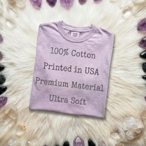 May include: A lilac-colored t-shirt with the text "100% Cotton, Printed in USA, Premium Material, Ultra Soft." The shirt is folded and placed on a fluffy, white surface, surrounded by crystals.