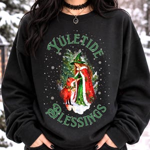 Op de afbeelding: Een zwart sweatshirt met lange mouwen en een grafische print van een persoon in een groene jurk en rode mantel met een groene hoed, staand bij een vos. De tekst YULETIDE BLESSINGS is in het groen boven en onder de afbeelding gedrukt.
