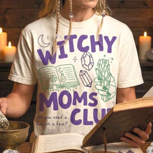 Op de afbeelding: Cr&egrave;mekleurig T-shirt met de woorden "Witchy Moms Club" in paars. Het shirt heeft groene en paarse illustraties van een boek, kristallen en een bundel kruiden. De tekst "do you need a hug or a hex?" staat ook op het shirt.