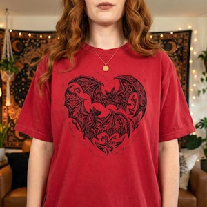 May include: Red t-shirt featuring a black heart-shaped design with bats. The graphic includes detailed wing patterns and decorative elements. The shirt has short sleeves and a crew neck.