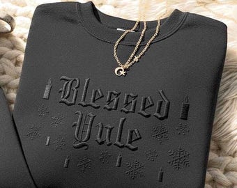 May include: A black crewneck sweatshirt featuring tonal black embroidery that reads Blessed Yule in a gothic font. The design includes embroidered candle and snowflake motifs surrounding the text. A delicate gold-tone chain necklace with a crescent moon charm rests on the neckline.