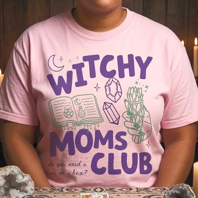 Op de afbeelding: Lichtroze t-shirt met de tekst "Witchy Moms Club" in paars. Het ontwerp bevat illustraties van een spreukenboek, kristallen en een saliebundel. De zin "do you need a hug or a hex?" staat er ook op.