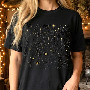 Könnte beinhalten: Schwarzes T-Shirt mit einem goldenen Sternen- und Sternbild-Design. Das Design zeigt Sterne und Linien in verschiedenen Größen, wodurch ein Nachthimmel-Effekt entsteht. Das Shirt besteht aus einem weichen Material.