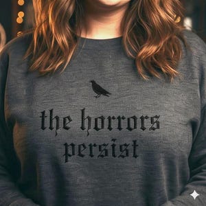 May include: Dark gray sweatshirt with the text "the horrors persist" in a gothic font. A black bird graphic is above the text. The sweatshirt is a crew neck style.