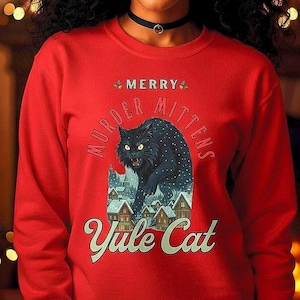 May include: A bright red long-sleeve sweatshirt featuring a graphic of a black cat with yellow eyes standing over a snowy village. The text reads MERRY MURDER MITTENS Yule Cat in a vintage-style font. The design includes small holly sprigs.