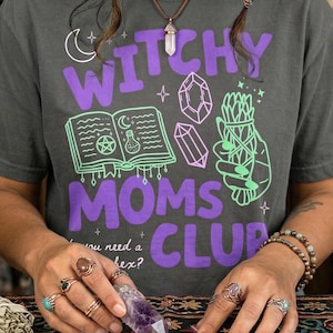 Op de afbeelding: Donkergrijs t-shirt met de tekst "Witchy Moms Club" in paars. Het shirt heeft groene en paarse afbeeldingen van een boek, kristallen en een bundel kruiden. De persoon draagt ringen en houdt een kristal vast.