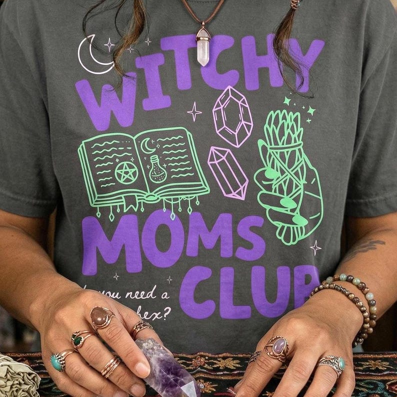 Op de afbeelding: Donkergrijs t-shirt met de tekst "Witchy Moms Club" in paars. Het shirt heeft groene en paarse afbeeldingen van een boek, kristallen en een bundel kruiden. De persoon draagt ringen en houdt een kristal vast.