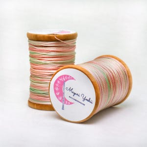 May include: Two spools of thread with a wooden base. The thread is a mix of pink, green, and white. One spool is upright, and the other is on its side. The label on the side of the spool reads "Mayuri Yoshii MAYUBOEHOUSE".