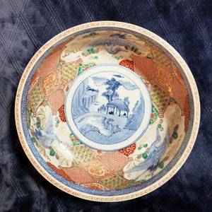 May include: A large, round, white ceramic bowl with a blue and red floral design. The bowl has a raised rim and a scalloped edge. The design features a central scene of a small house in a landscape, surrounded by stylized flowers and leaves. The bowl is decorated with gold accents.