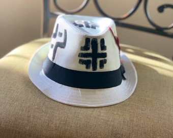 May include: A white fedora hat with a black band and black geometric designs on the crown.