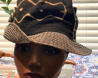 May include: A black hat with a tan patterned design and a striped brim. The hat is displayed on a mannequin head. The hat's design features a repeating wave pattern.