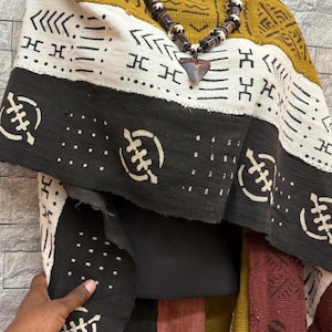 Peut inclure: Un châle en tissu à motifs noir, blanc et jaune moutarde, drapé sur un mannequin. Un collier de perles avec un pendentif triangulaire est porté sur le châle. Le tissu présente des motifs géométriques et symboliques.