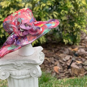 May include: A large, colorful hat with a pink flower and a white base. The hat is made of a sheer fabric with a floral pattern. The hat is sitting on a white pedestal.
