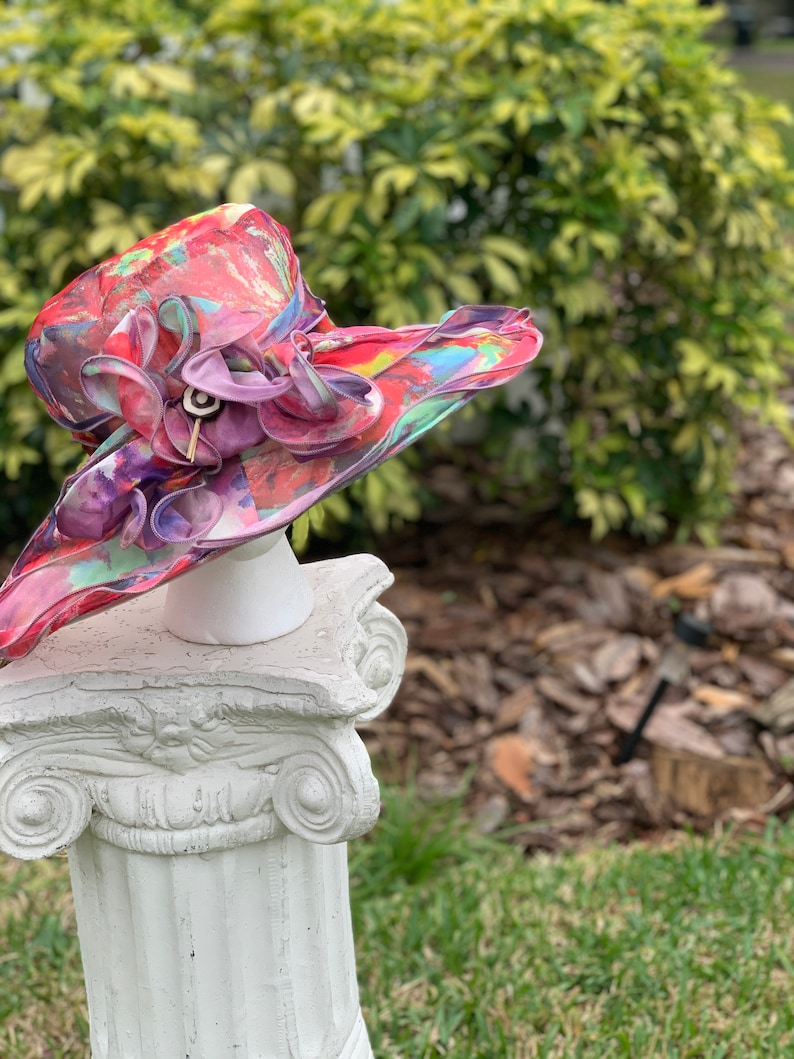 May include: A large, colorful hat with a pink flower and a white base. The hat is made of a sheer fabric with a floral pattern. The hat is sitting on a white pedestal.