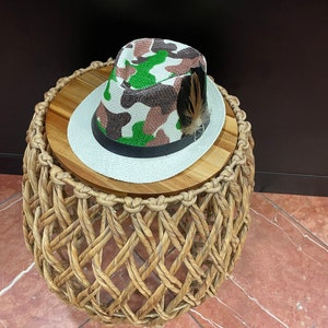 May include: A camouflage fedora hat with a black band and a feather accent sits on a woven wicker stool with a wooden top.